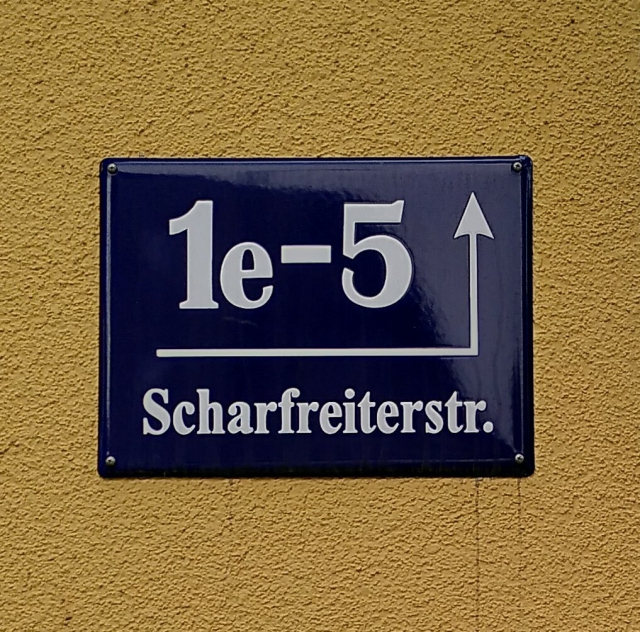A blue house number sign with white text, mounted on a yellow wall. The sign reads "1e-5, Scharfreiterstraße".