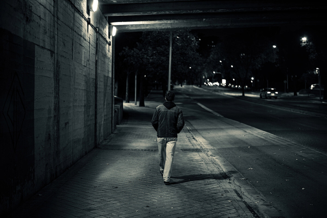 Fotografía nocturna en blanco y negro de una acera y calzada bajo un puente levemente iluminado. Se ve un chico vestido con cazadora y pantalones vaqueros caminando yendo andando hacia al fondo.