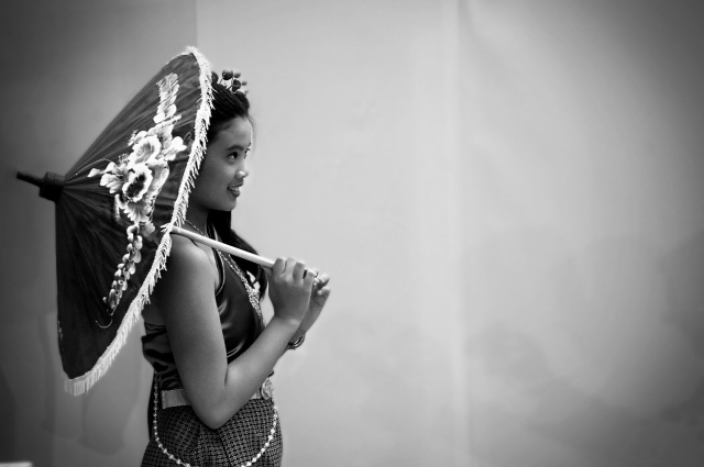 Foto en blanco y negro en plano americano de bailarina thai esperando para salir a realizar su número de baile (de perfil, mirando a la derecha) portando paraguas tradicional para danza apoyado en el hombro.