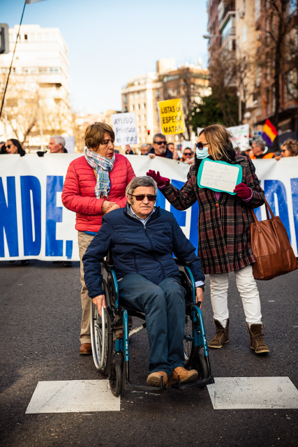 Retrato de un hombre en silla de ruedas con dos acompañantes, delante de la cabecera, una de ellas con un cartel que indica “No puedo hablar, pero estoy aquí, 100% Sanidad Pública”. Columna este de la Manifestación por la Sanidad en Madrid que sale desde el Hospital de La Princesa.