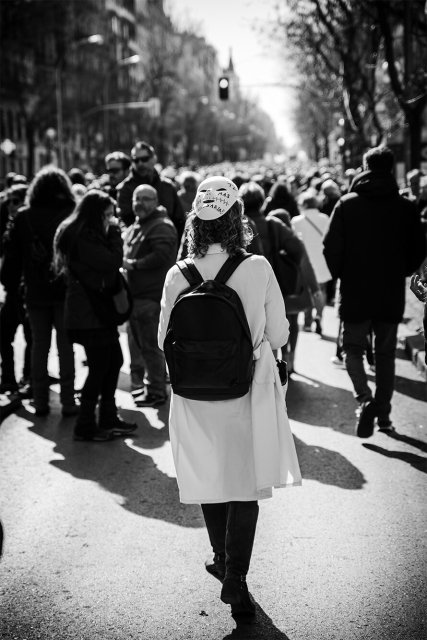 Foto en plano entero y vertical de una de las manifestantes sanitarias con una mascara clásica de protesta en la parte trasera de su cabeza. Se ve de fondo y desenfocado la columna de manifestantes hasta el final de la calle. Columna este de la Manifestación por la Sanidad en Madrid que sale desde el Hospital de La Princesa. 