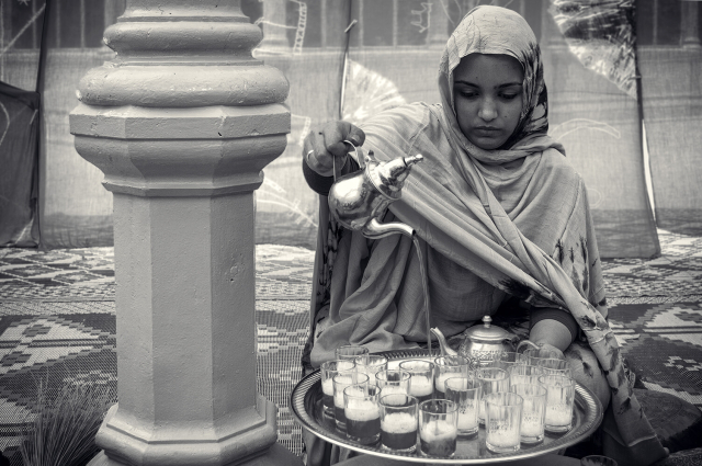 Retrato blanco y negro y en plano medio de una chica saharaui sentada en el suelo demostrando su habilidad para servir el té ceremonial sobre una bandeja con multitud de vasos. Va vestida con una melhfa tradicional (en color, de verde) y está sentada en una alfromba dentro de una jaima, al fondo se pueden ver las telas que dan al exterior.