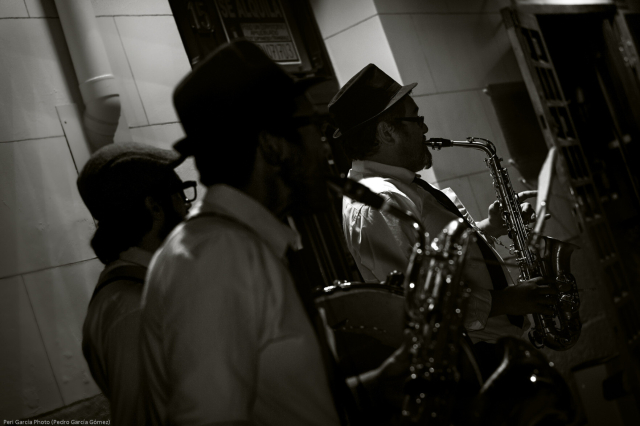 Foto de perfil en plano medio y en blanco y negro de un trío de músicos (dos saxofonistas y un tamborilero, todos con camisa, corbata y sombrero) tocando Jazz en la calle en una noche de verano madrileña, los dos más próximos a cámara están desenfocados con el otro (saxofonista) enfocado al fondo y recibiendo la luz de una farola.