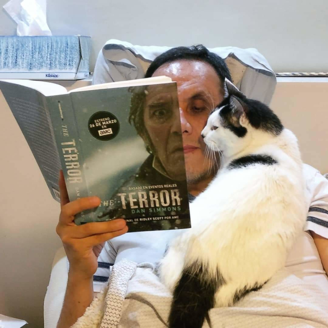 Un hombre lee "El Terror" de Dan Simmons y su cara completa la que se ve en la portada del libro. En su hombro, un gato parece leer también.