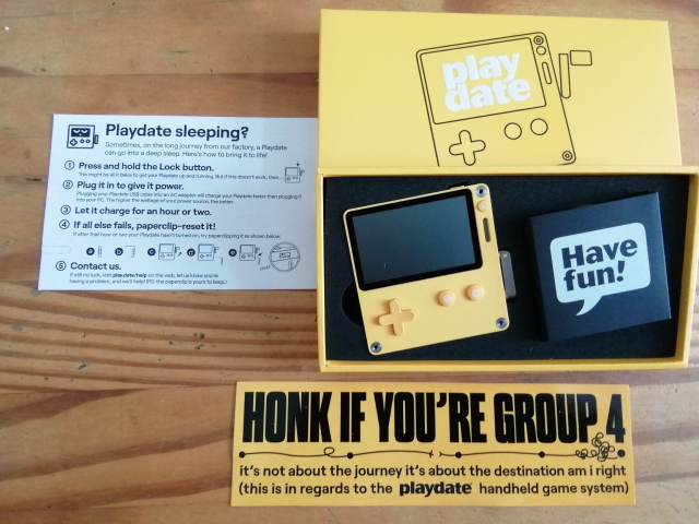 Picture of the PlayDate handheld game console with it's original packaging and a Sticker saying "Honk if you're group 4" next to it.