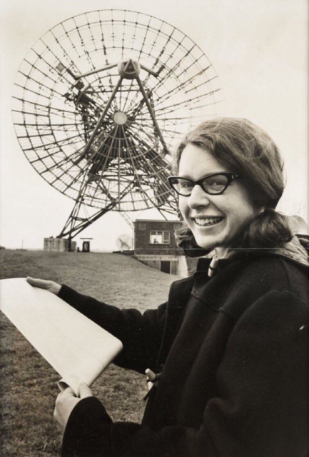 Black and white photo of Jocelyn Bell Burrell, when she was a graduate student, holding a printout on a roll of paper and standing in front of a large radio dish. She is wearing a coat and glasses, smiling and looking toward the camera.