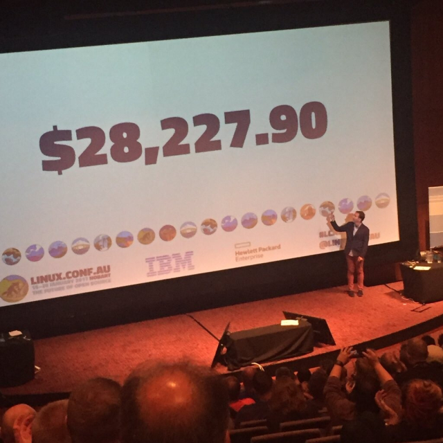 Chris stands at the front of the stage at linux.conf.au 2017, there's a giant slide behind him that says "$28,227.90".