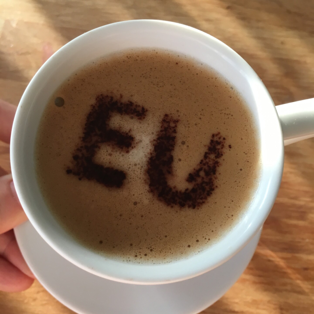 A close-up photo of a cup of coffee from above. The text “EU” is created inside the cup by playing with the lather.