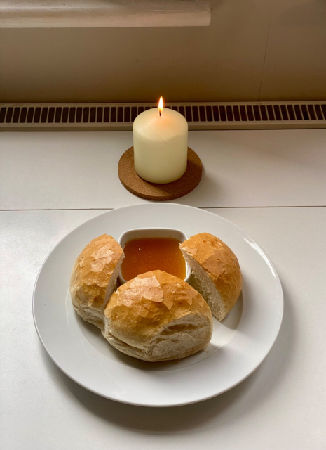 A white circular plate on a white tabletop. On the plate is a large bread roll cut in three sections and a small square dish of honey. A wide candle stands lit on a cork mat behind the plate 