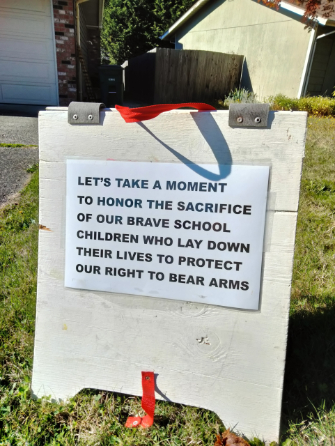 Sign reading "LET'S TAKE A MOMENT TO HONOR THE SACRIFICE OF OUR BRAVE SCHOOL CHILDREN WHO LAY DOWN THEIR LIVES TO PROTECT OUR RIGHT TO BEAR ARMS"