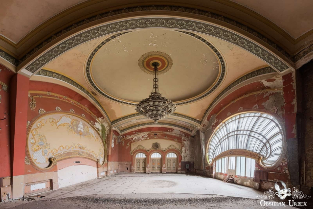 art nouveau casino, the room is empty. the architecture is stunning with red walls. there is a chandelier and large shell shaped windows