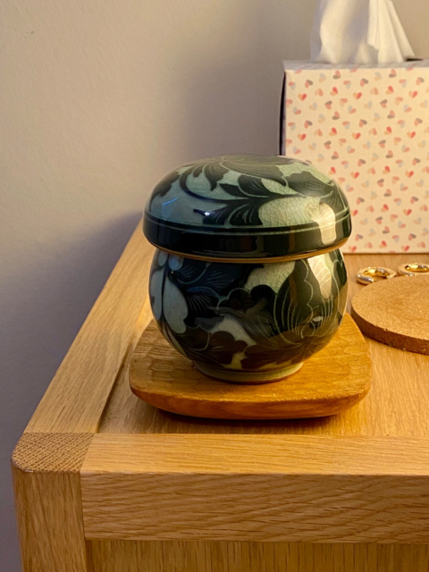 A fat and rounded Korean ceramic teacup with a wide round lid that makes it look a bit like a mushroom, sitting on my pale oak bedside cabinet. It’s glazed in a pattern of flowing dark green foliage on a light green background 