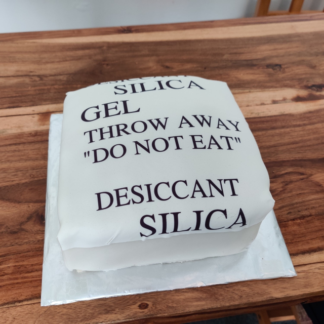 a square cake with white icing and the words "Desiccant Silica Gel. Throw away. Do Not Eat" printed on it.