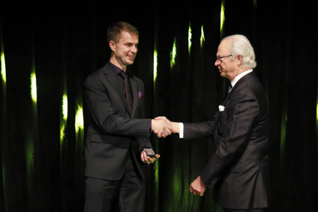 Daniel shakes hands with the king of Sweden on a dark, greenish background. Both are wearing suits.