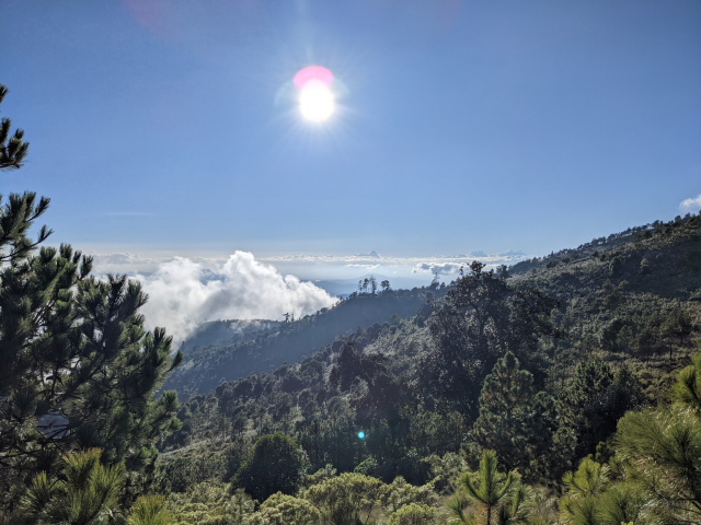 el sol del atardecer visto desde la laguna. En el cielo ni una nube pero en el horizonte, abajo, se alza alguna. Cubriendo la tierra hay muchos pinos y pastizales.