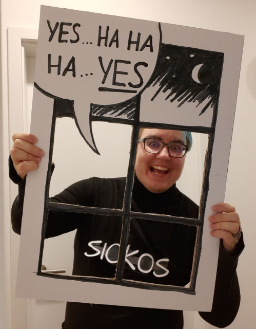 Ein weißer trans Mann mit blauen Haaren hat sich als das "Sickos"-Meme verkleidet mit dunkel geschminkten buschigen Augenbrauen, einem schwarzen Pullover, auf dem "Sickos" steht, und einem gebastelten Fensterrahmen mit der Sprechblase "Yes ... HA HA HA ... YES"