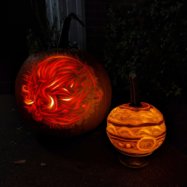Two carved pumpkins, lit from within, photographed at night. The larger pumpkin is carved with a detailed, stylized image of the sun including prominences that arc off the limb of the sun. The smaller pumpkin is carved like a planet, resembling a stylized Jupiter.