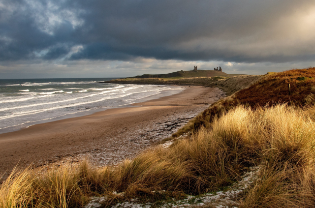 A photo of a beach with a light covering of snow. In the foreground sand dunes grasses are lit up with sunlight. Further back white water waves make their way up the beach. On the horizon the ruins of a castle can be seen under dark moody clouds. 