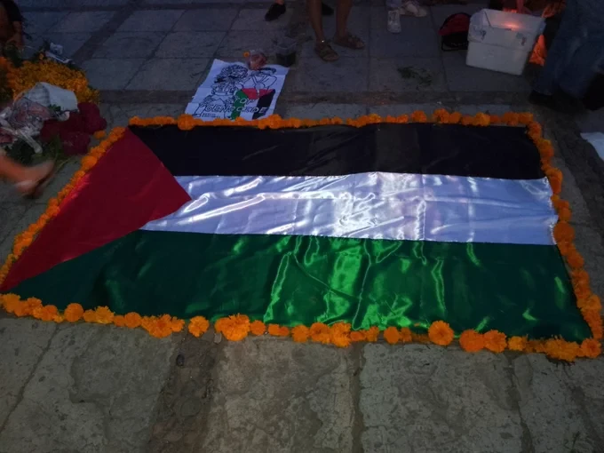 Una bandera de Palestina (triángulo rojo con una base en el astabandera y tres franjas horizontales: negra, blanca y verde) rodeada de cempaxóchitl, veladoras y dibujos de los asesinados.