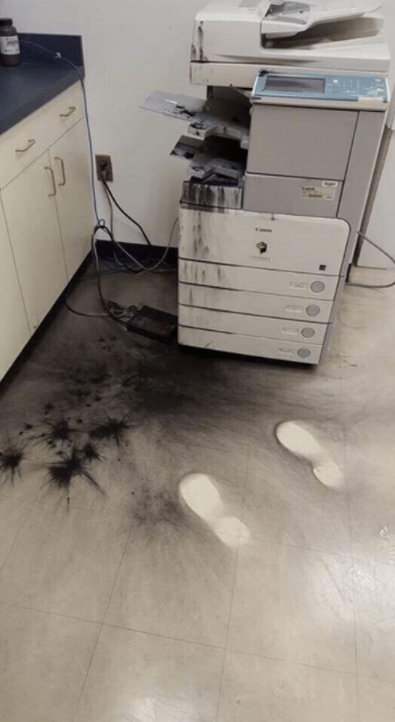 A picture of an ink cartridge for an office printer that has exploded in front of someone, and there is ink everywhere. What is notable are a set of footprints that keep the floor clean, almost as if the blast of the ink exploding has missed it.