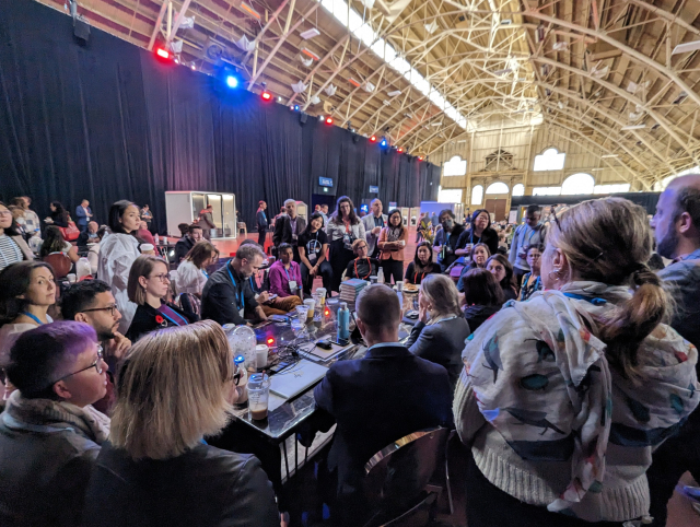 Group of people sitting and standing around a table in a very large room with arched ceiling.  