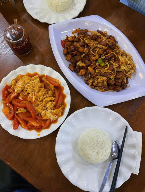 Wooden table with 2 plates of rice, a plate of Tomato Fried Eggs with generous amounts of tomatoes and its juices, and a big serving of Spicy Chicken which also comes with flat noodles. A container of Chilli Oil is on the side.