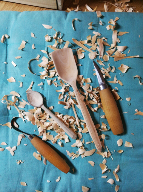 Two wooden spoons, a hook knife and a carving knife on top of a turquoise cushion sprinkeled with wood shavings
