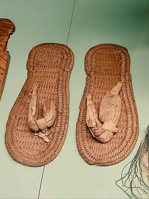 The picture shows a completely preserved pair of Egyptian sandals,  they resemble modern flip flops. They are made of woven reed and palm leaves, the sole with bound edges.
