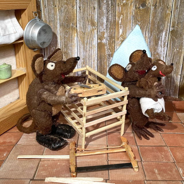 Photo of Minimus and Minima, the Latin mice, with the baby mouse. Minimus is working on a wooden cradle with a plane, while a bow saw is lying next to him. Minima is holding the baby in her paws