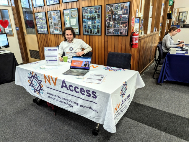 Sean sitting at a table with an NV Access tablecloth, brochures and a laptop debating NVDA