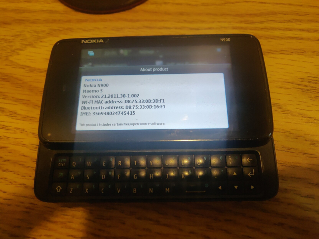 An old smartphone in landscape mode, with the keyboard slid out. on the display is the "about phone" info screen, showing that this is a Nokia N900, running Maemo 5, version 21.2011.38-1.002, plus mac addresses and IEMI number