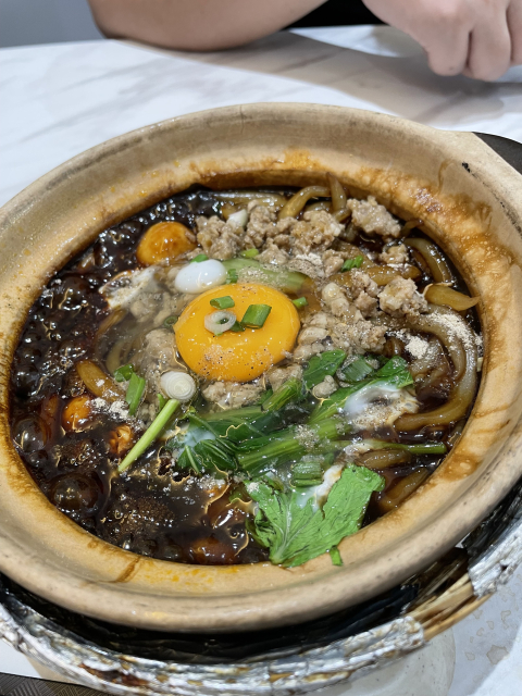 A hot claypot filled with thin rice noodles, flavorful broth, minced meat, veggies, and a raw egg with the runny egg yolk floating on top.