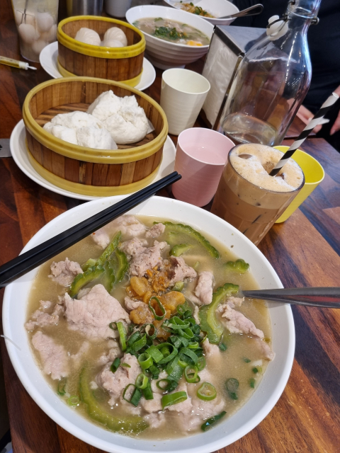 Table of food: noodles in rich pork broth topped with sliced meat, minced meat, bittergourd slices, spring onions, fried lard. Also, ice milk tea, prawn dumplings, meat buns, lychee drink