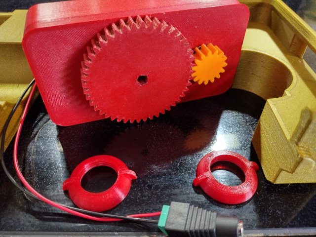 Several parts of a motorized burr coffee grinder sit on the black CERAN surface of a stove showing two halves of a 3D printed clamp housing, two 3D printed wing nuts, and a 3D printed motor housing. This time, the motor housing is tipped on its side to expose two 3D printed gears, the larger of which has a 5-sided hole into which the top of the hand grinder is to be inserted.