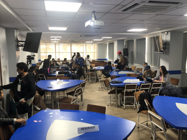 People in a conference room. There are triangular tables in the front, and rows of chairs in the back.