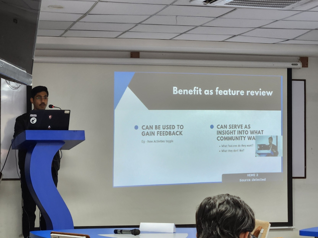 A South Asian man stands up on a stage, in front of a podium. A projector screen shows the slides of his presentation. The slide is titled, "Benefit as feature review".