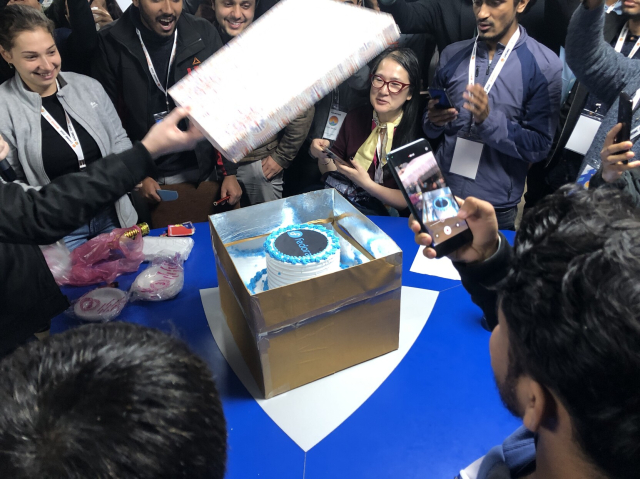 White cake with a black and blue top, inside a box. The box's lid is being removed while people watch intently.