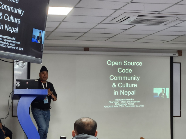 A South Asian male stands at a speaker podium on a stage. A projector screen with a slide show presentation is next to him. The slide shown is the title slide, "Open Source Code, Community, & Culture in Nepal".