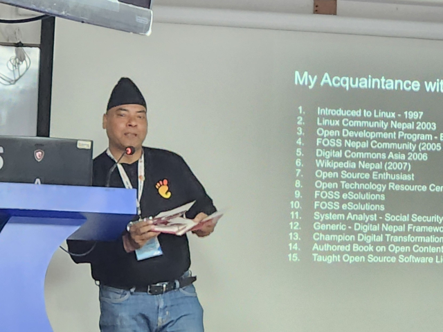 A South Asian male stands at a speaker podium on a stage. A projector screen with a slide show presentation is next to him. He is holding a CD booklet with a copy of the first Nepali Linux distribution from the early 2000s.