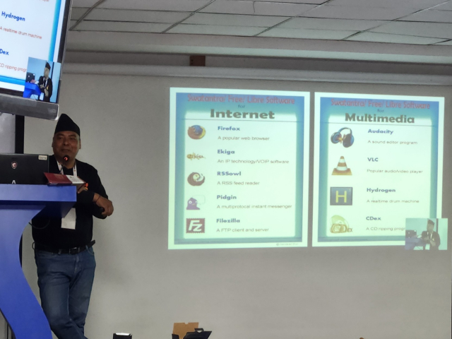 A South Asian male stands at a speaker podium on a stage. A projector screen with a slide show presentation is next to him. The slide shown lists common Open Source tools for Internet browsing and multimedia tools.
