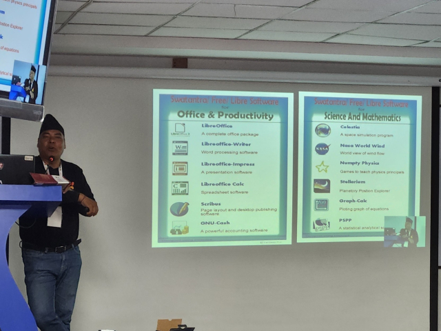 A South Asian male stands at a speaker podium on a stage. A projector screen with a slide show presentation is next to him. The slide shown lists Open Source tools for office and productivity tools, and science and mathematics applications.