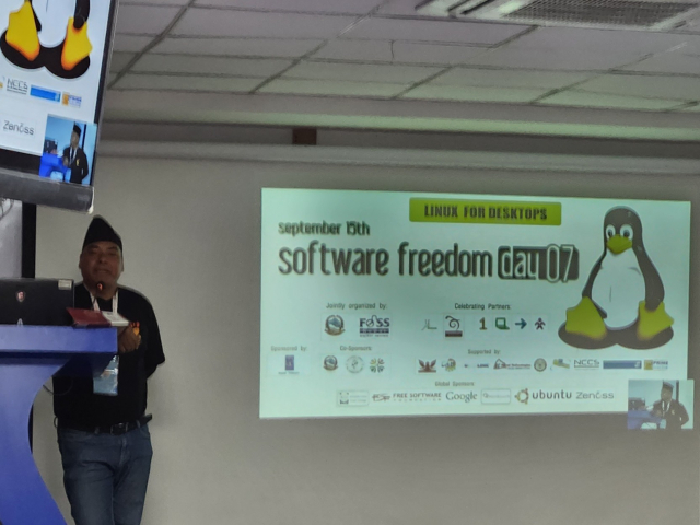 A South Asian male stands at a speaker podium on a stage. A projector screen with a slide show presentation is next to him. The slide shown describes a local Software Freedom Event from long ago in Nepal.
