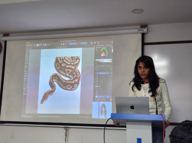 A South Asian woman stands at a speaker podium in front of a laptop. A projector screen with a slide show is next to her. The slide shown is a window from Krita with a snake, showing the workflow for creating new art.