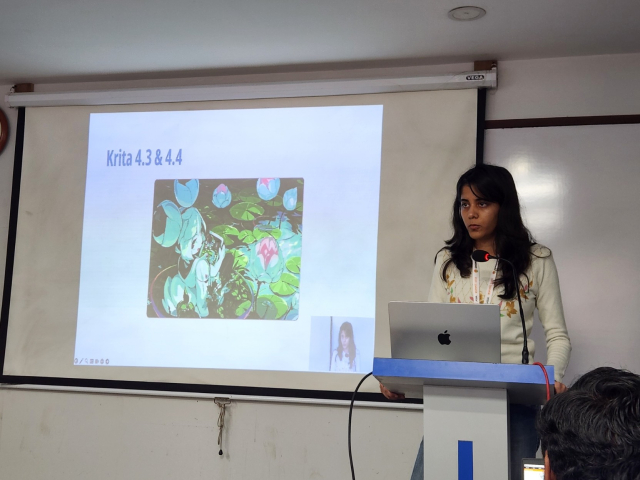 A South Asian woman stands at a speaker podium in front of a laptop. A projector screen with a slide show is next to her. The slide shown is highlighting the Kriti 4.3 and 4.4 versions.