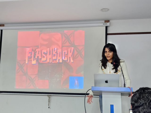 A South Asian woman stands at a speaker podium in front of a laptop. A projector screen with a slide show is next to her. The slide shown is with large text reading, "Flashback".