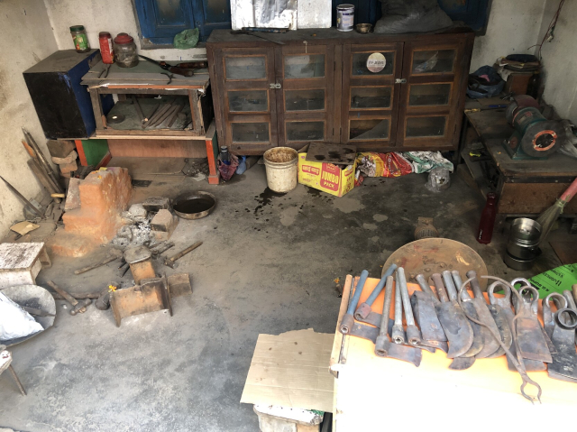 A tiny blacksmith's shopfront. There's a small charcoal forge on the floor, a small anvil from a railroad tie, hammers, tongs. The tools on display are axes, knives, spades, and calipers of some sort. There is a grinding wheel on a desk.