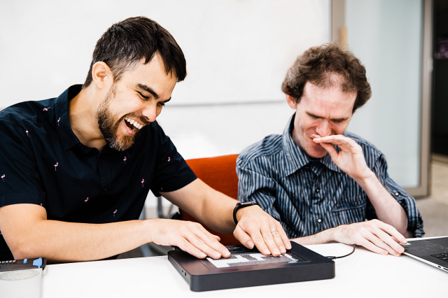 Jamie and Mick smiling and laughing, working with a Braille tablet