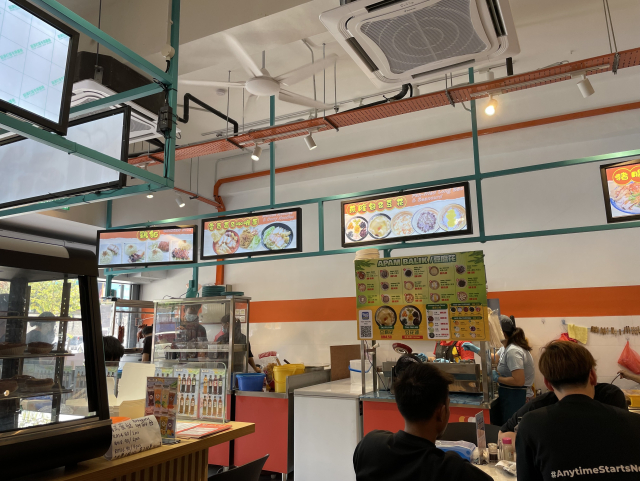 The interiors of a shop filled with customers enjoying food around round tables and with dozens of food stalls tended by individual hawkers selling different types of food