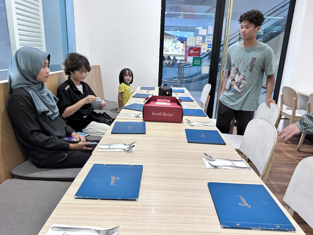 A group of people at a restaurant table with menus, a standing boy looking towards the camera, and a seated woman and children.