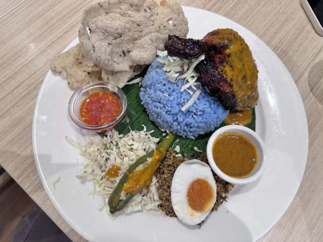 A plate of Nasi Kerabu with blue rice, grilled chicken percik, crackers, boiled egg, salad, and sauces.